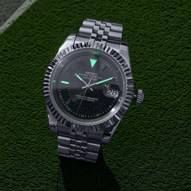 Low light top view of Seiko Mod Datejust Wimbledon with glowing green markers and brushed steel bracelet visible.