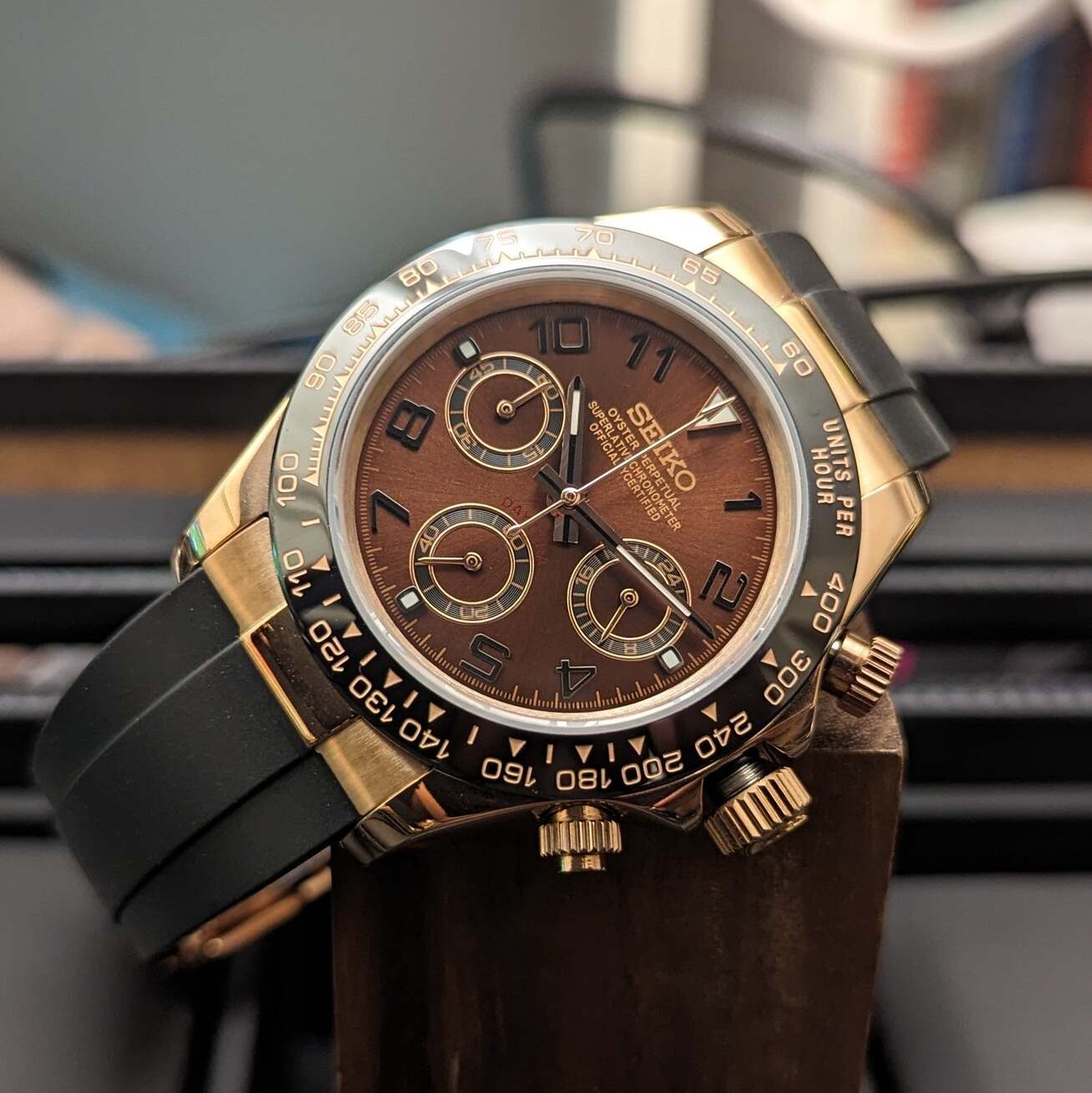 Front left angle of a rose gold Seiko Mod Daytona with brown dial, black Arabic numerals, three subdials, and black rubber strap resting on a wooden stand.