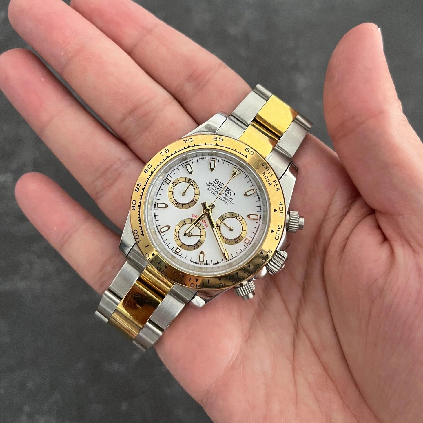Two tone Seiko Mod Daytona with gold bezel and white dial, held in a hand against a dark background.