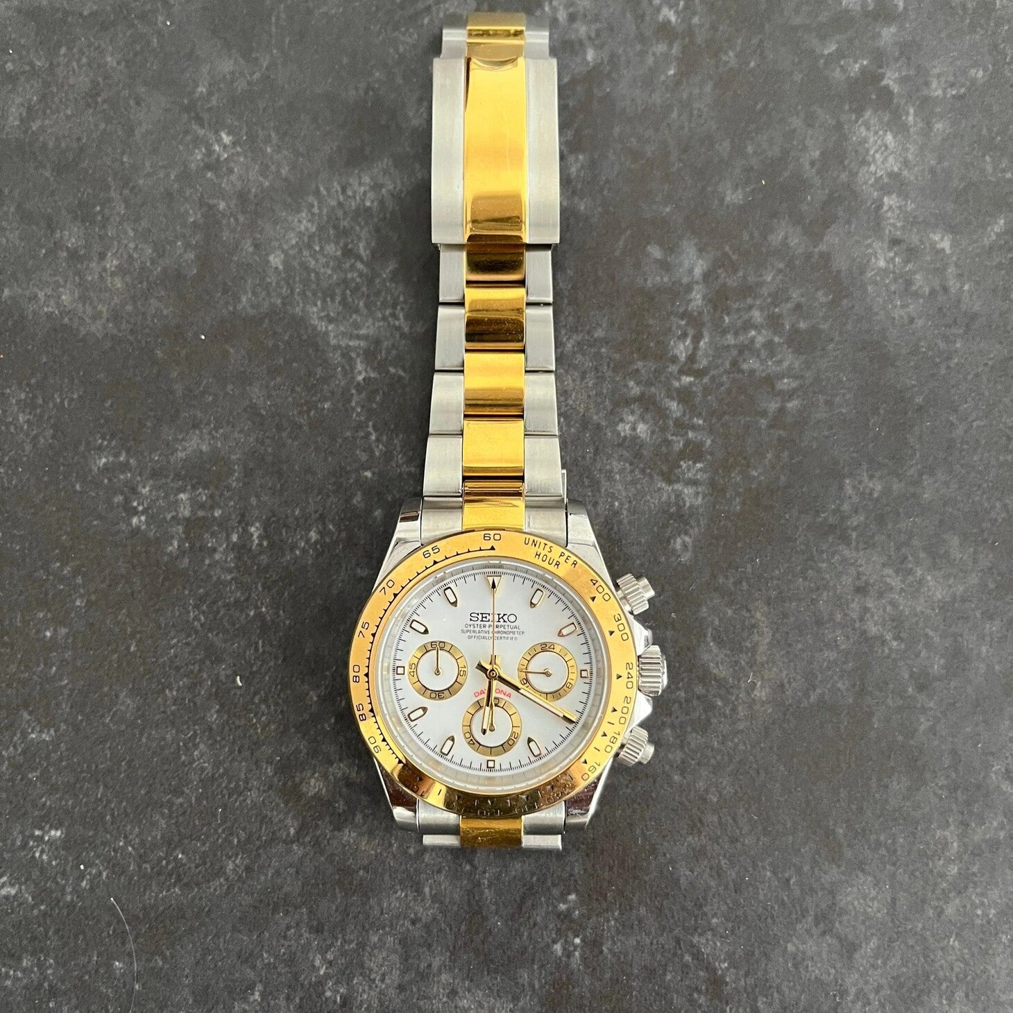 Flat lay of Seiko Mod Daytona two tone gold and silver bracelet with white dial, placed on a textured grey surface.