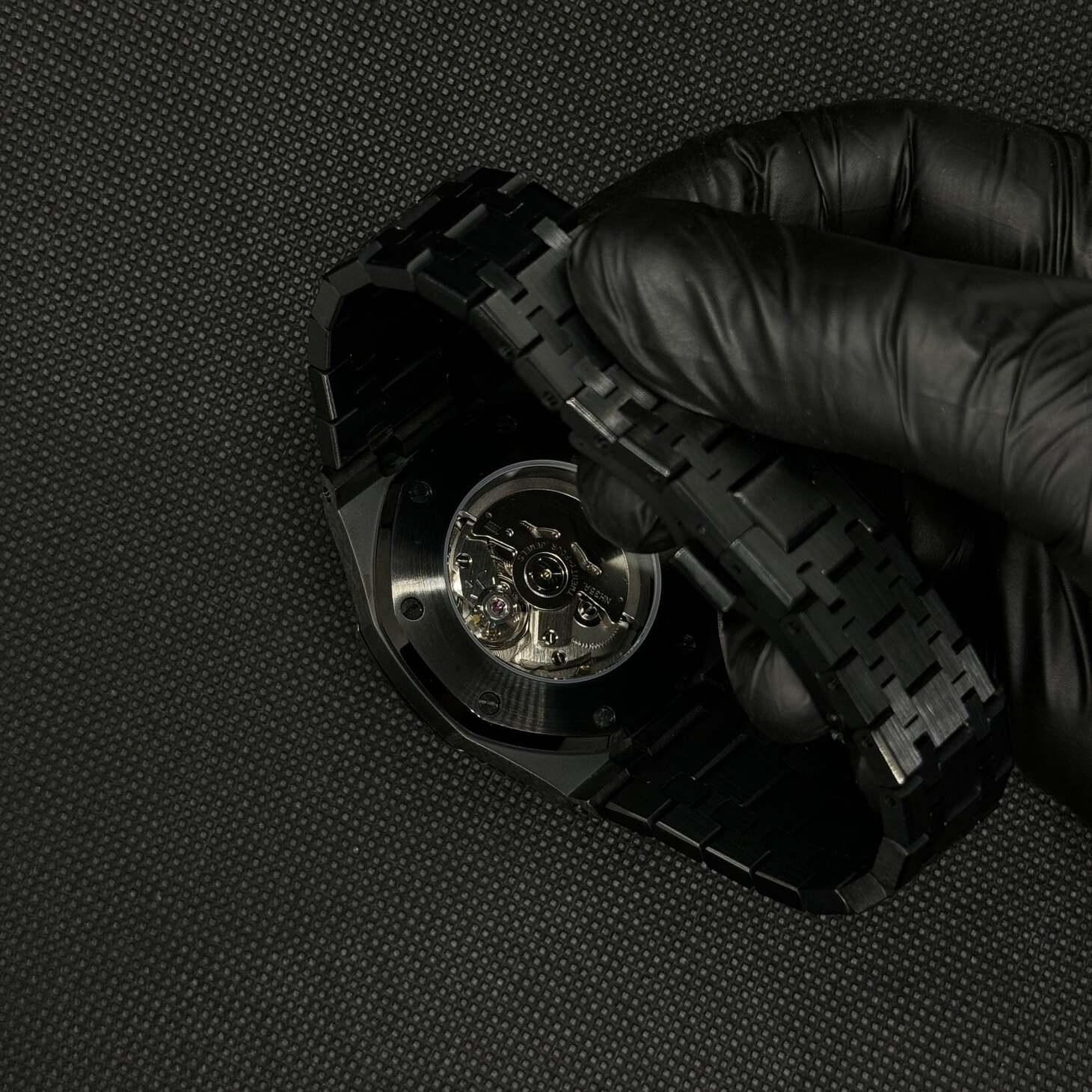 Bottom view of the Seiko Mod Royal Oak automatic watch in all black, showing the open crystal case back with visible Seiko NH35 movement and brushed stainless steel bracelet held by a gloved hand