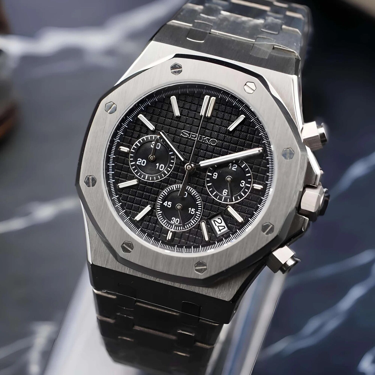 Side angled close up of the Seiko Mod Royal Oak VK63 chronograph watch with black textured dial, three subdials, brushed stainless steel case, steel bracelet, and date window at 4 o’clock