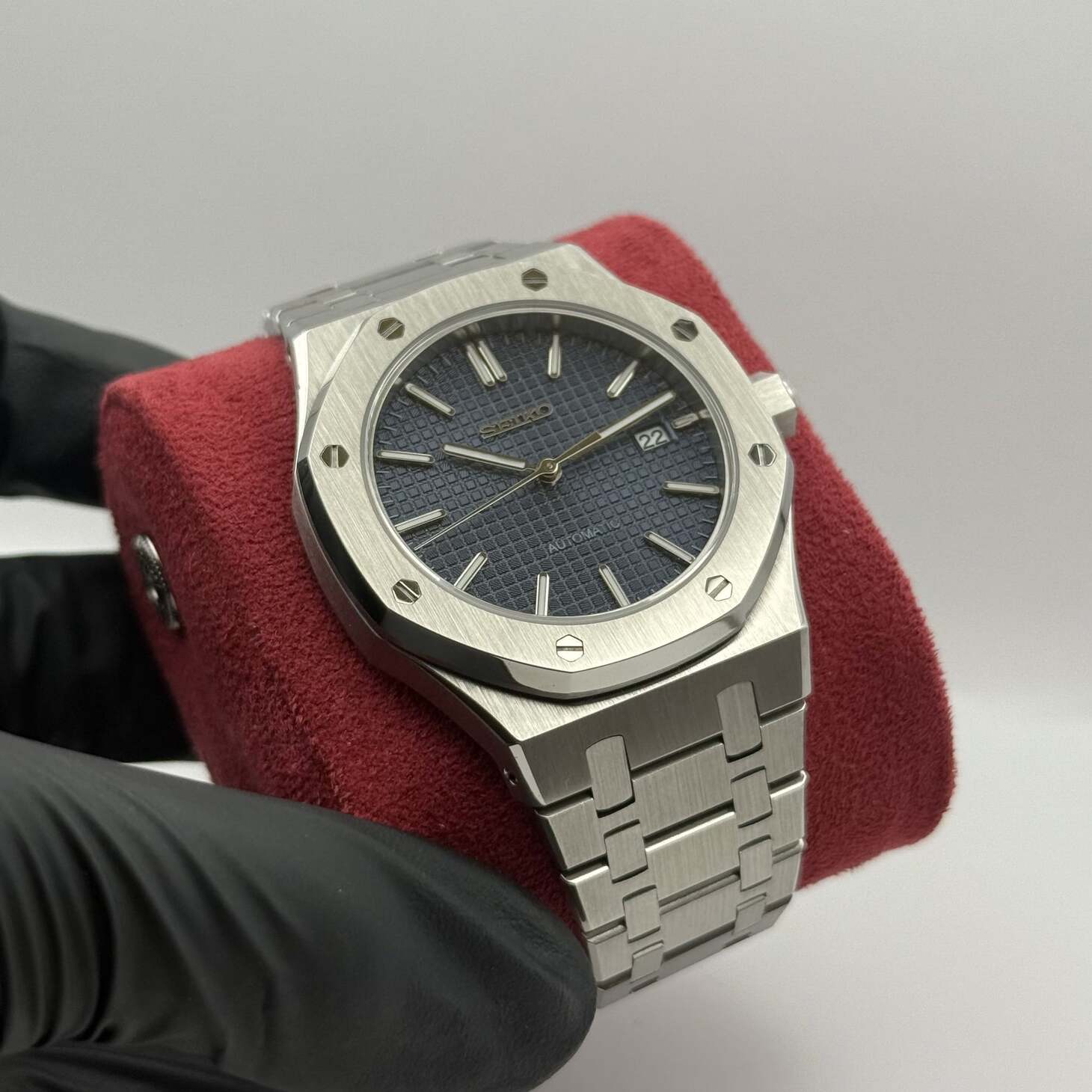 Angled side view of the Seiko Mod Royal Oak automatic watch with navy blue textured dial, stainless steel bracelet, and date at 3 o’clock placed on a red cushion held by a gloved hand