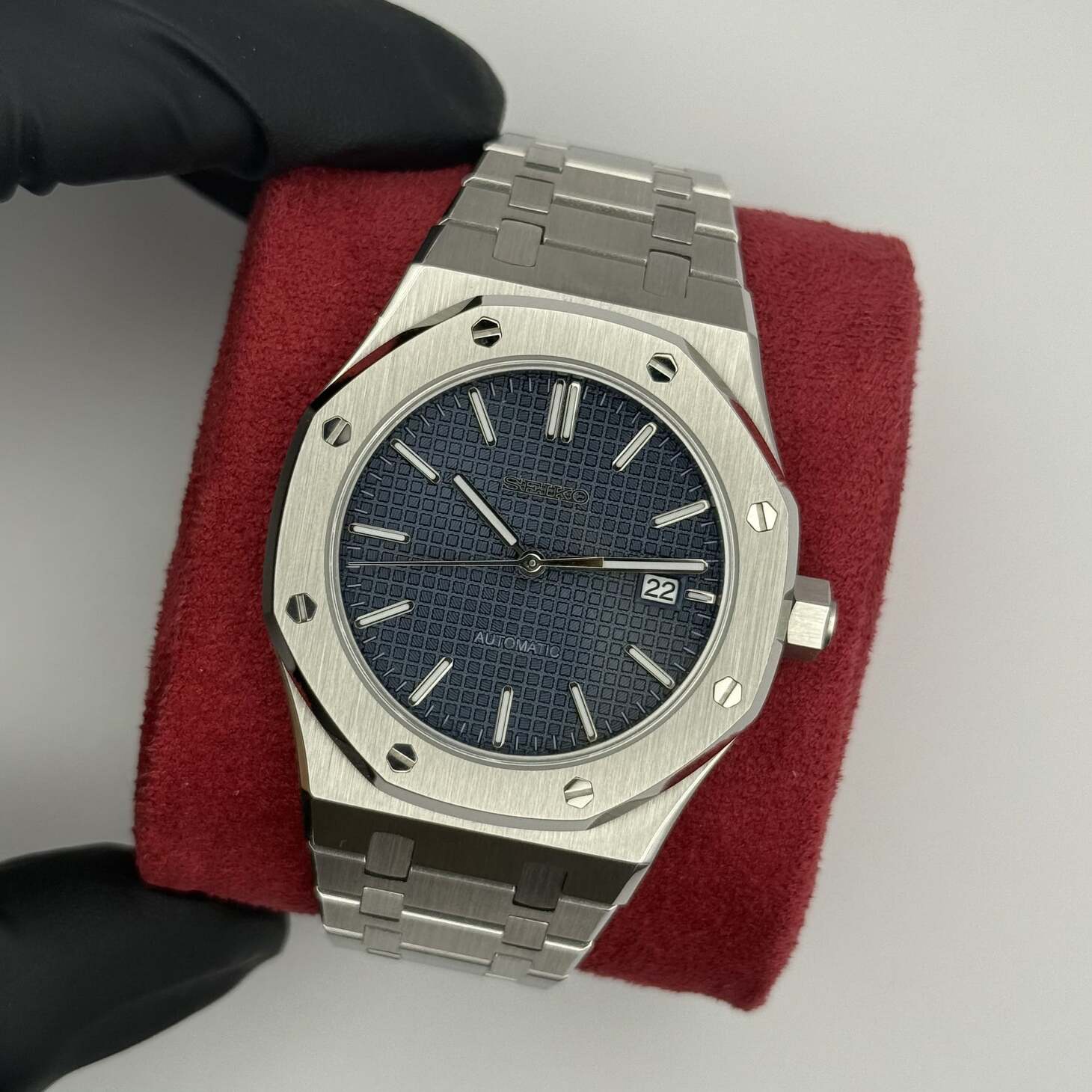 Top down view of the Seiko Mod Royal Oak automatic watch with navy blue grid pattern dial, stainless steel bracelet, and date display at 3 o’clock resting on a red cushion