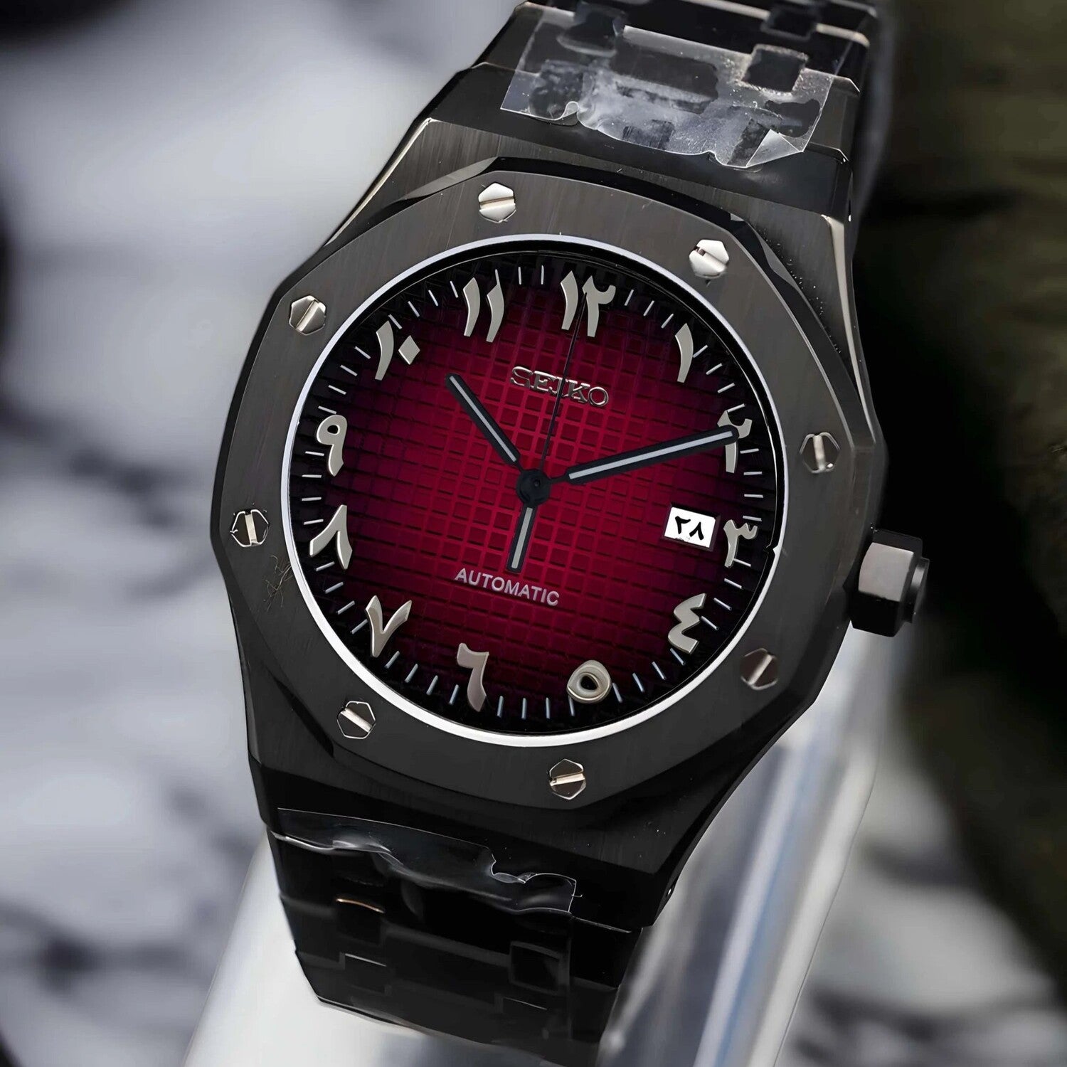 Right angled side view of a Seiko Mod Royal Oak watch with a striking red grid textured dial, silver Arabic numerals, and bold black hands, set in a brushed black stainless steel case with exposed screws on the octagonal bezel and matching bracelet.