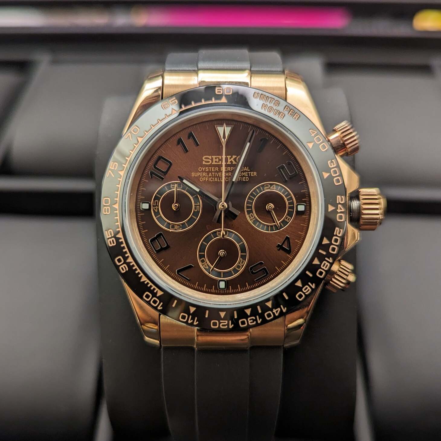 Straight on view of a rose gold Seiko Mod Daytona with sunburst brown dial, Arabic numerals, three subdials, and black rubber strap, placed on a display stand.